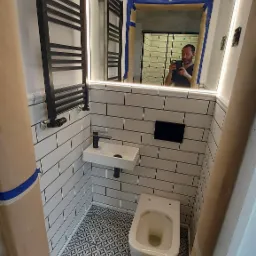 Wąska łazienka w trakcie prac wykończeniowych z białymi kaflami, wzorzystą podłogą, małą umywalką, wiszącą toaletą i czarnym grzejnikiem drabinkowym; w lustrze odbija się osoba robiąca zdjęcie.