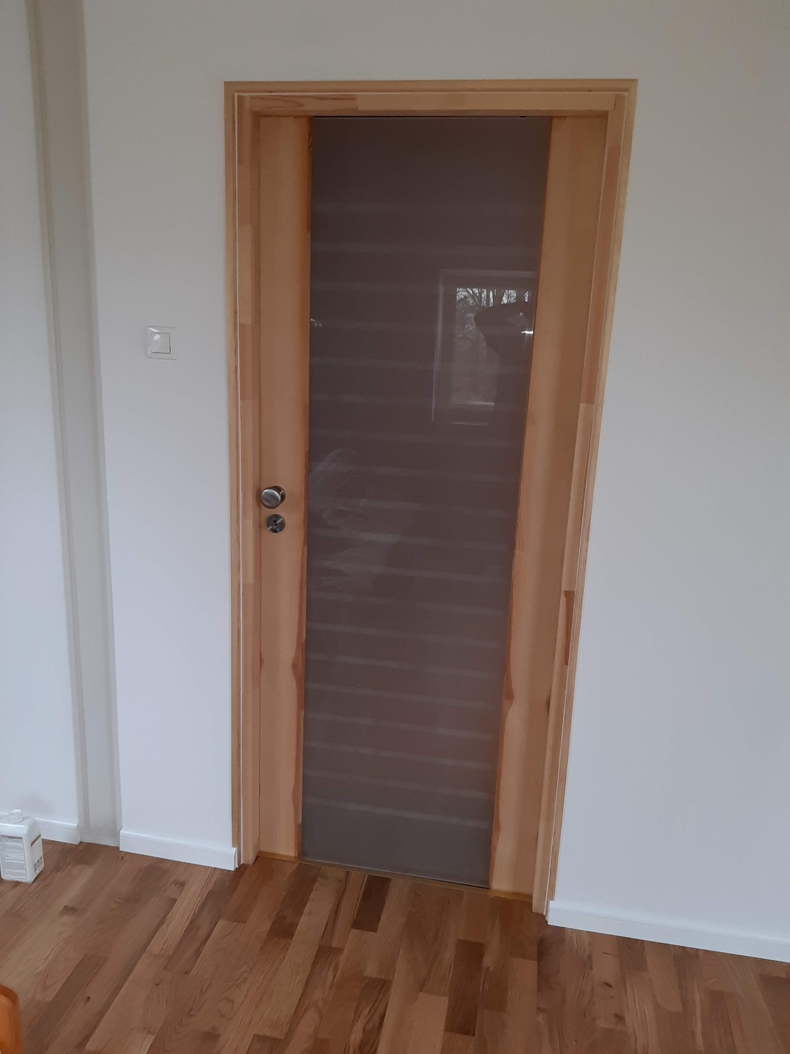 Drewniane drzwi wewnętrzne z matową szybą, zamontowane w pomalowanym na biało pomieszczeniu z drewnianą podłogą, widoczny włącznik światła i listwa przypodłogowa.