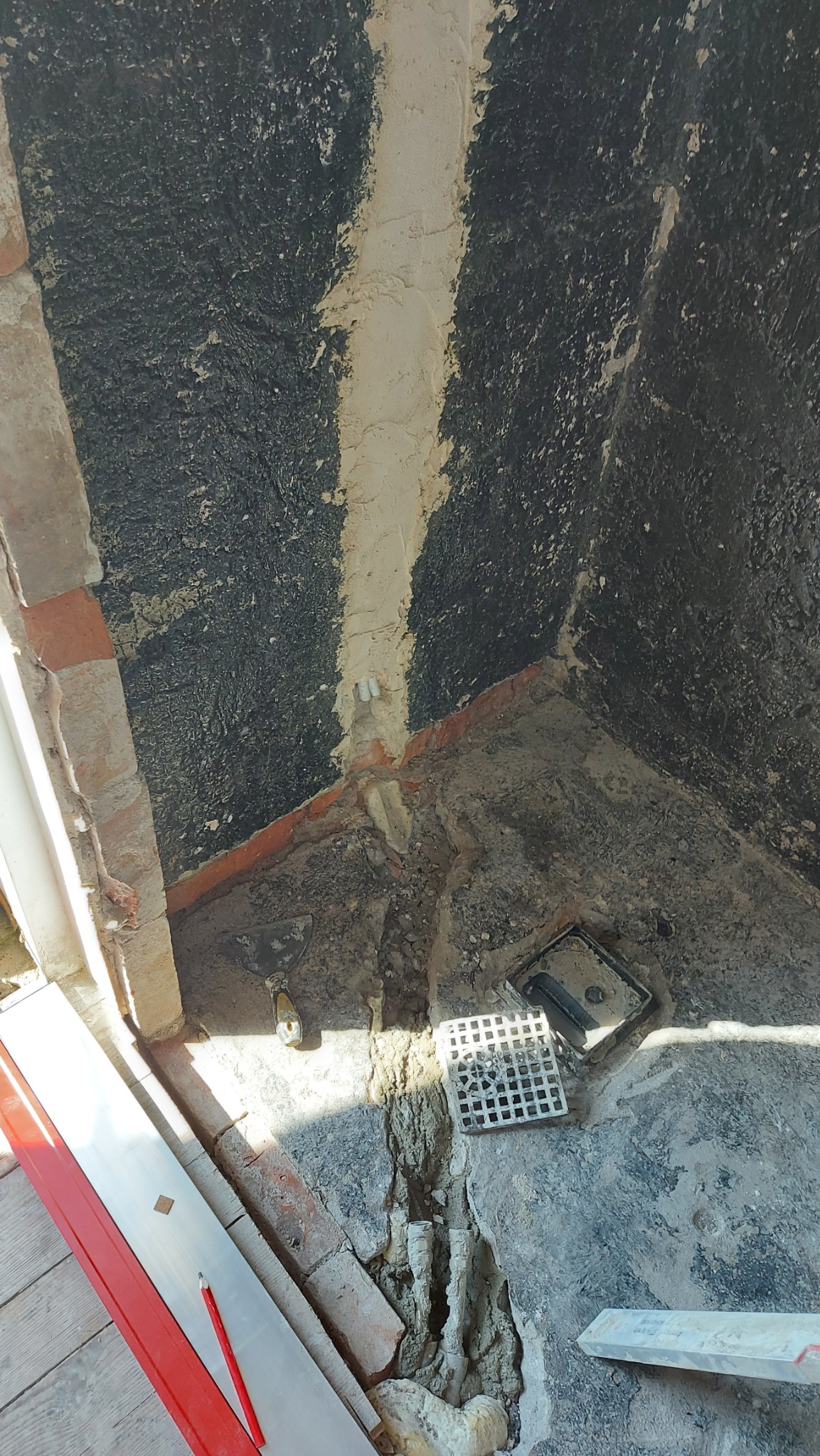 Przygotowanie do remontu łazienki: surowe ściany z nałożonym gruntem, niezabezpieczone rury, kratka odpływowa, poziomica i szpachelka w rogu pomieszczenia.