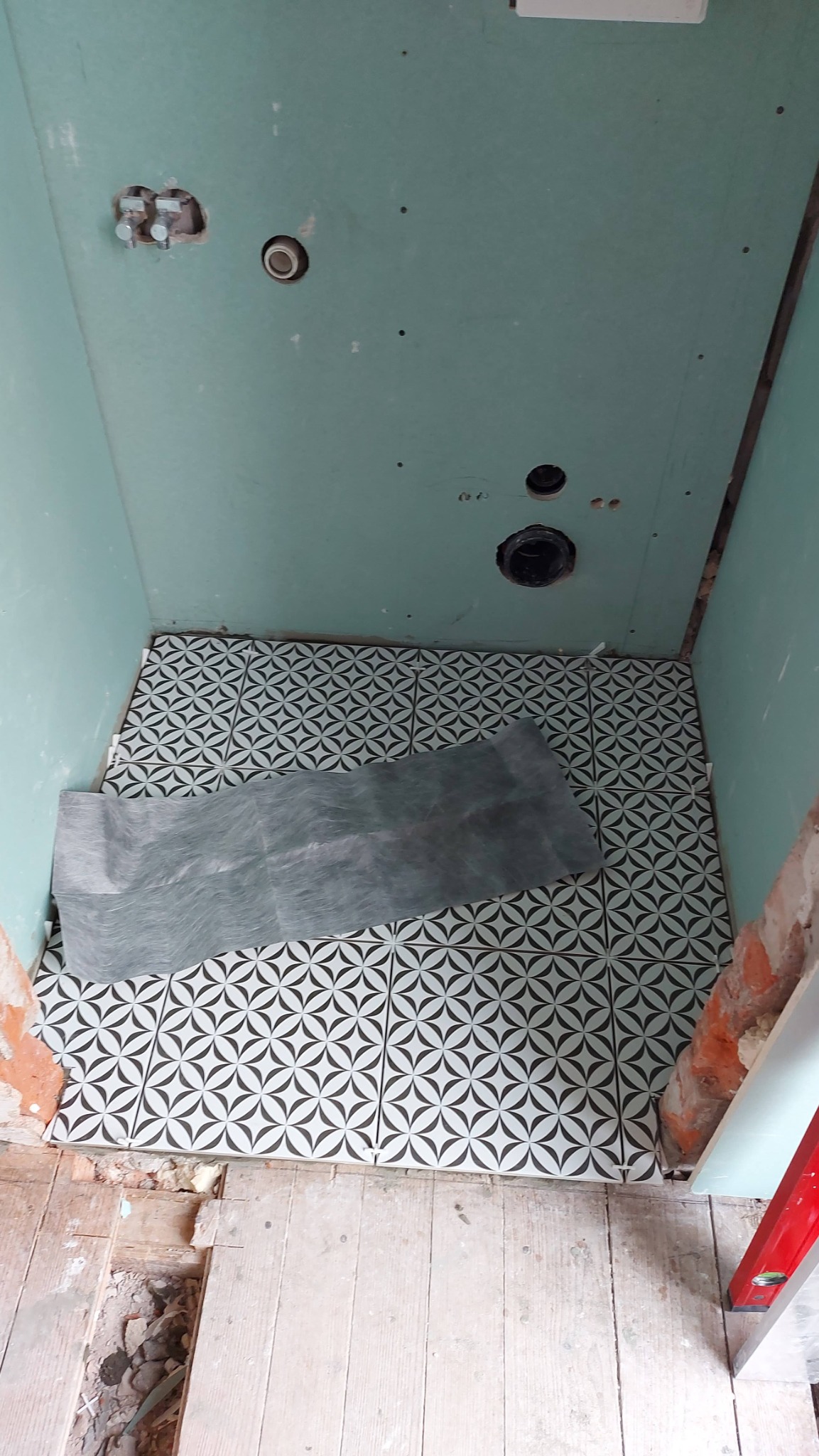 Fragment łazienki w trakcie układania wzorzystych, czarno-białych płytek podłogowych; widoczne ściany z płyt kartonowo-gipsowych z otworami na instalacje, fragment starej drewnianej podłogi...