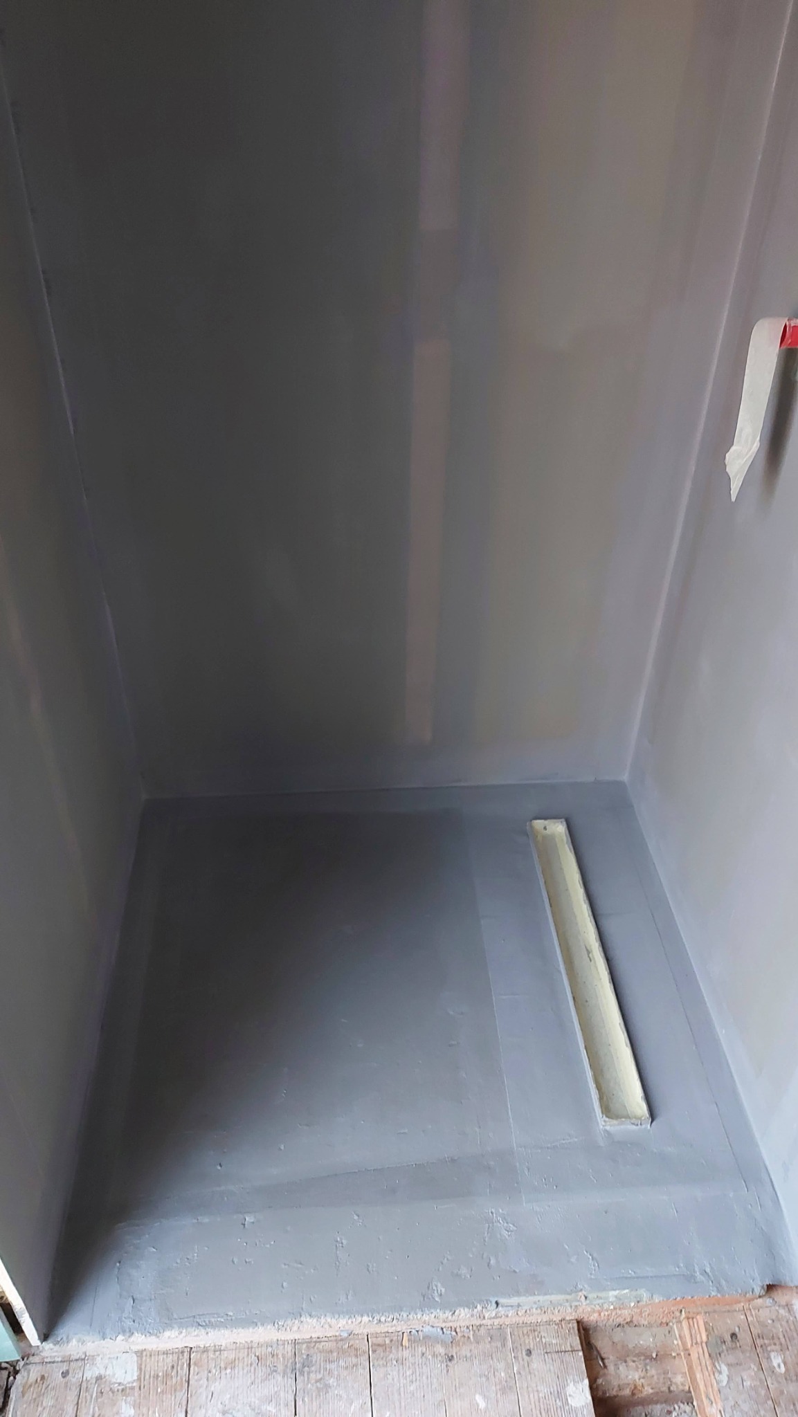 Przygotowana do hydroizolacji szara kabina prysznicowa z odpływem liniowym, zabezpieczona taśmą malarską na łączeniach ścian i podłogi, widoczne surowe deski podłogowe poza obszarem kabiny.