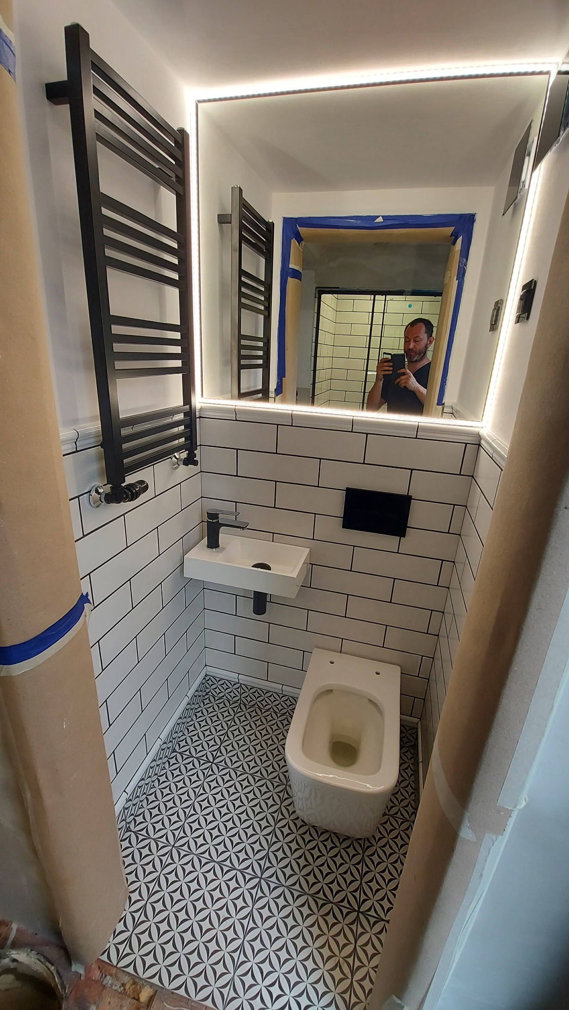 Wąska łazienka w trakcie prac wykończeniowych z białymi kaflami, wzorzystą podłogą, małą umywalką, wiszącą toaletą i czarnym grzejnikiem drabinkowym; w lustrze odbija się osoba robiąca zdjęcie.