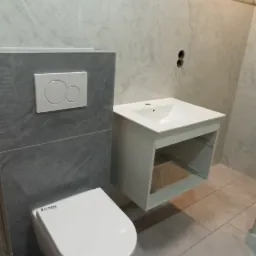 Wisząca toaleta i umywalka z szafką w surowym, wykafelkowanym wnętrzu łazienki z widocznymi kablami elektrycznymi w ścianie.