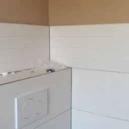 Świeżo zamontowany, podtynkowy stelaż WC z białym przyciskiem spłukującym na tle białej boazerii i beżowej ściany, widoczne ślady po montażu i karton po opakowaniu.