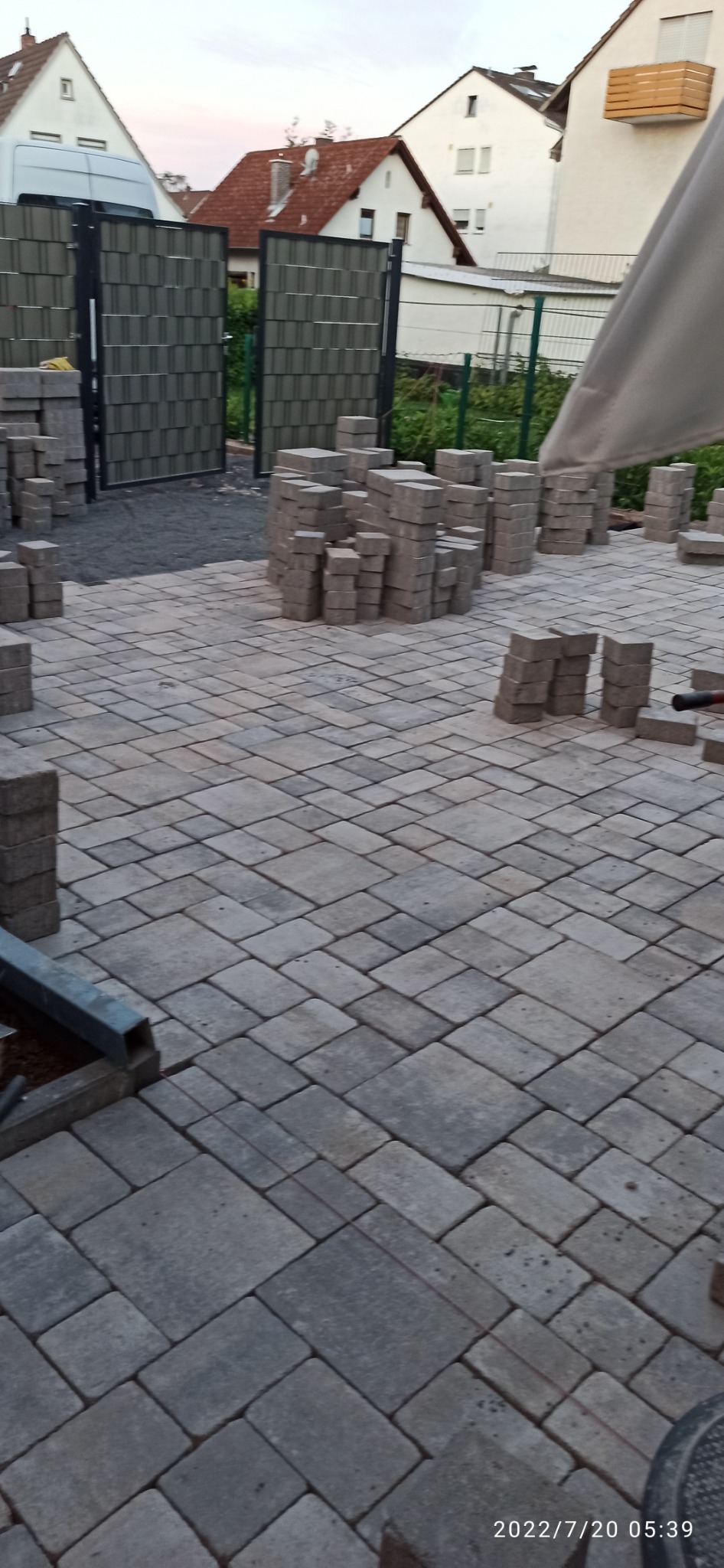 Układanie szarej kostki brukowej na posesji, widoczne stosy kostki przygotowane do ułożenia, w tle ogrodzenie i zabudowania mieszkalne.