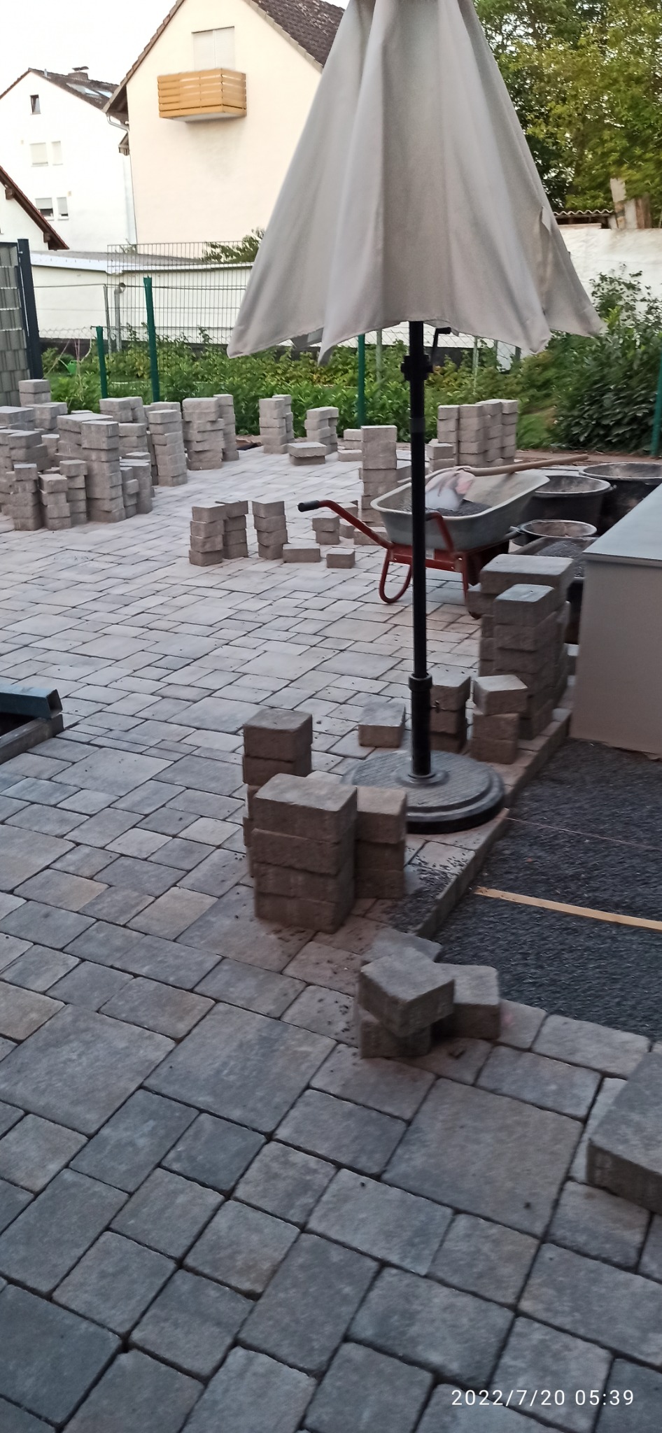 Układanie szarej kostki brukowej na posesji, widoczny parasol ogrodowy, taczka z narzędziami i przygotowane stosy kostki.