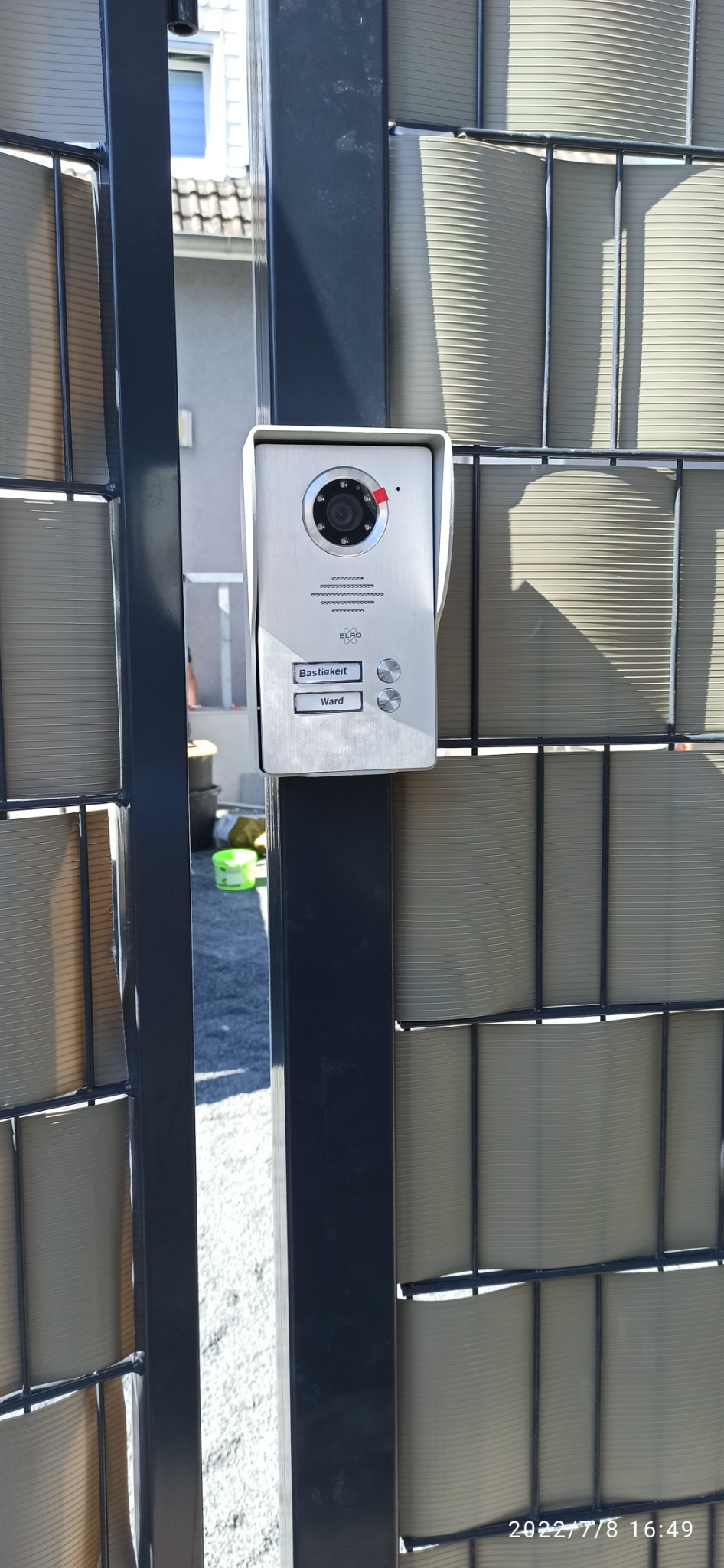 Srebrny wideodomofon ELRO z dwoma przyciskami (Bastigkeit, Ward) zamontowany na czarnym słupku ogrodzenia panelowego z szarym wypełnieniem, widok z bliska.