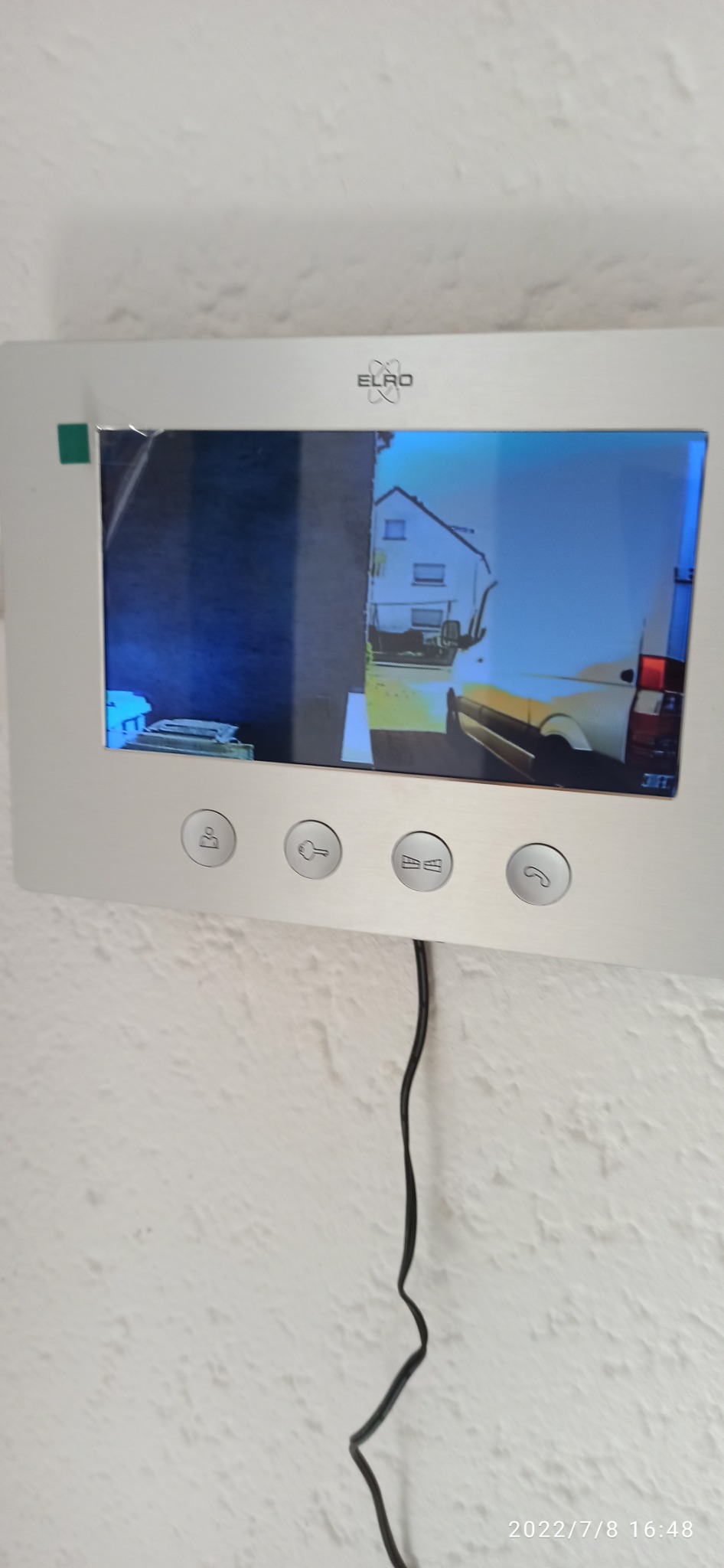Ekran domofonu ELRO zamontowanego na białej, teksturowanej ścianie, wyświetlający obraz z kamery na zewnątrz, gdzie widoczny jest budynek i zaparkowany biały samochód dostawczy.