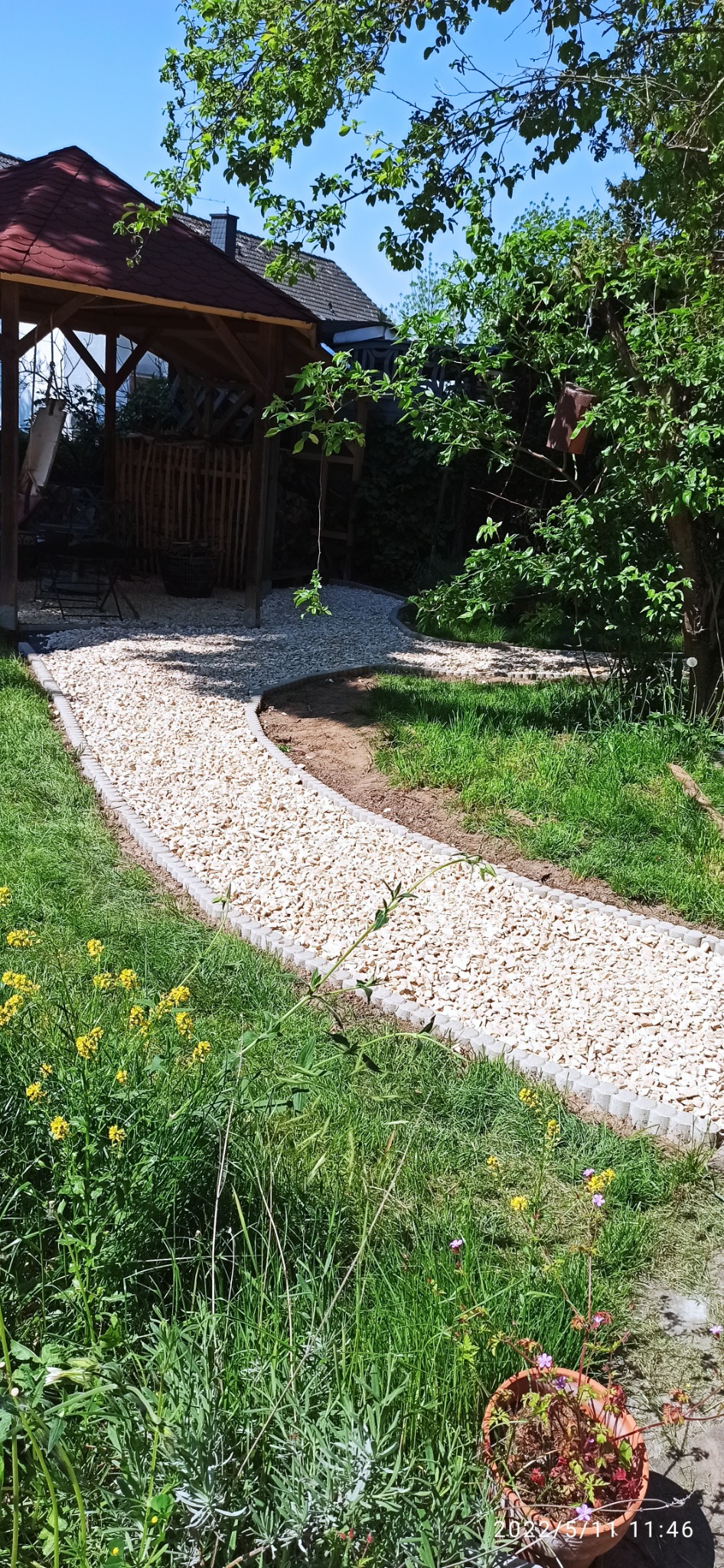 Kamyczkowa ścieżka ogrodowa z obrzeżami z kostki prowadząca do drewnianej altany, otoczona trawnikiem i kwitnącymi żółtymi kwiatami.