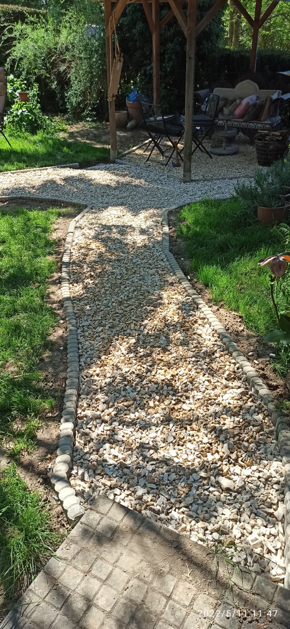 Kamykowa ścieżka ogrodowa z obrzeżami z betonowych kostek, prowadząca do drewnianej altany z meblami ogrodowymi w tle, otoczona trawnikiem i roślinnością.