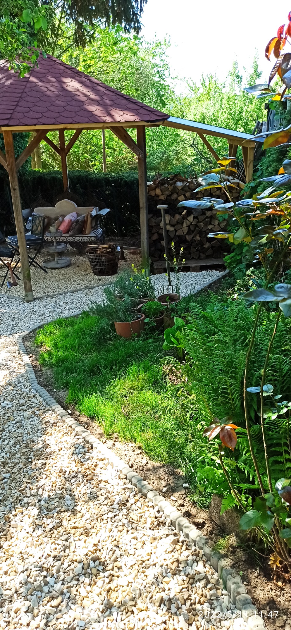 Altana ogrodowa z paleniskiem, drewnem opałowym i kamienną ścieżką, otoczona zielenią, w słoneczny dzień.