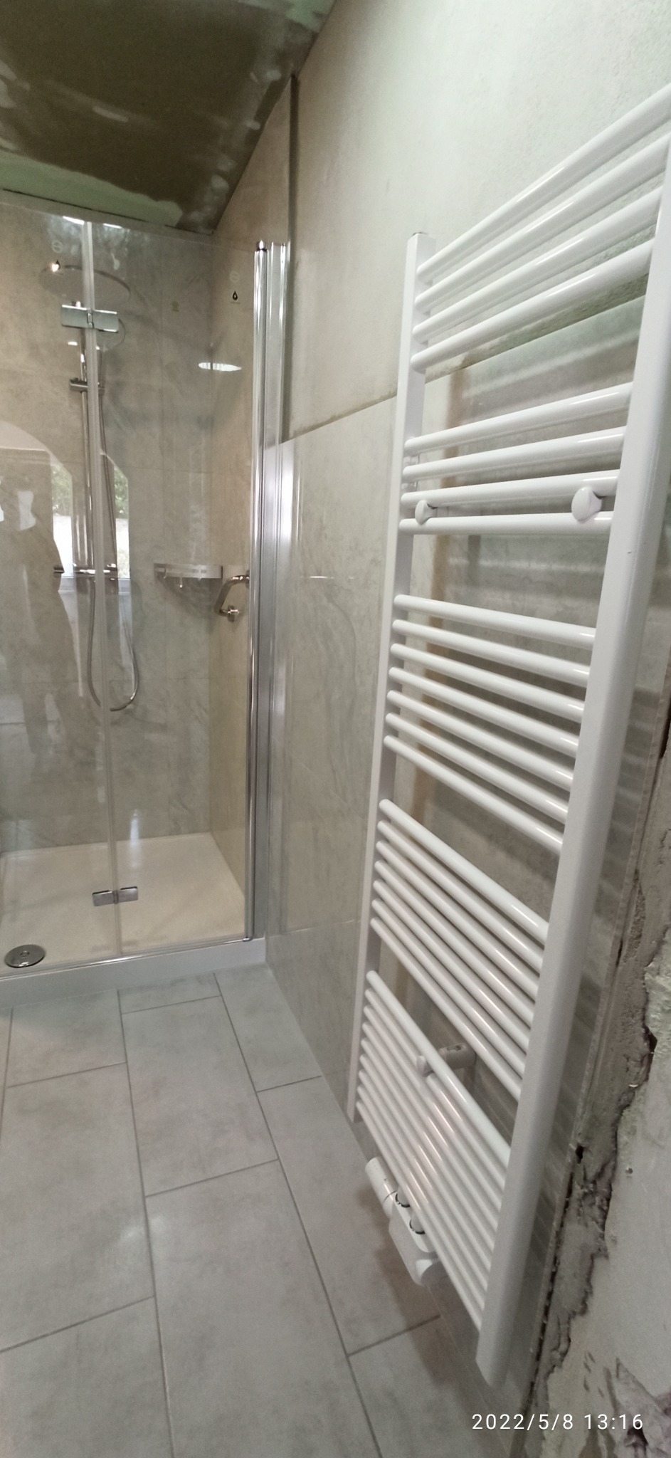 Nowoczesna łazienka z kabiną prysznicową z transparentnego szkła i białym grzejnikiem drabinkowym na tle ściany wyłożonej płytkami imitującymi marmur, widoczne pozostałości po remoncie.