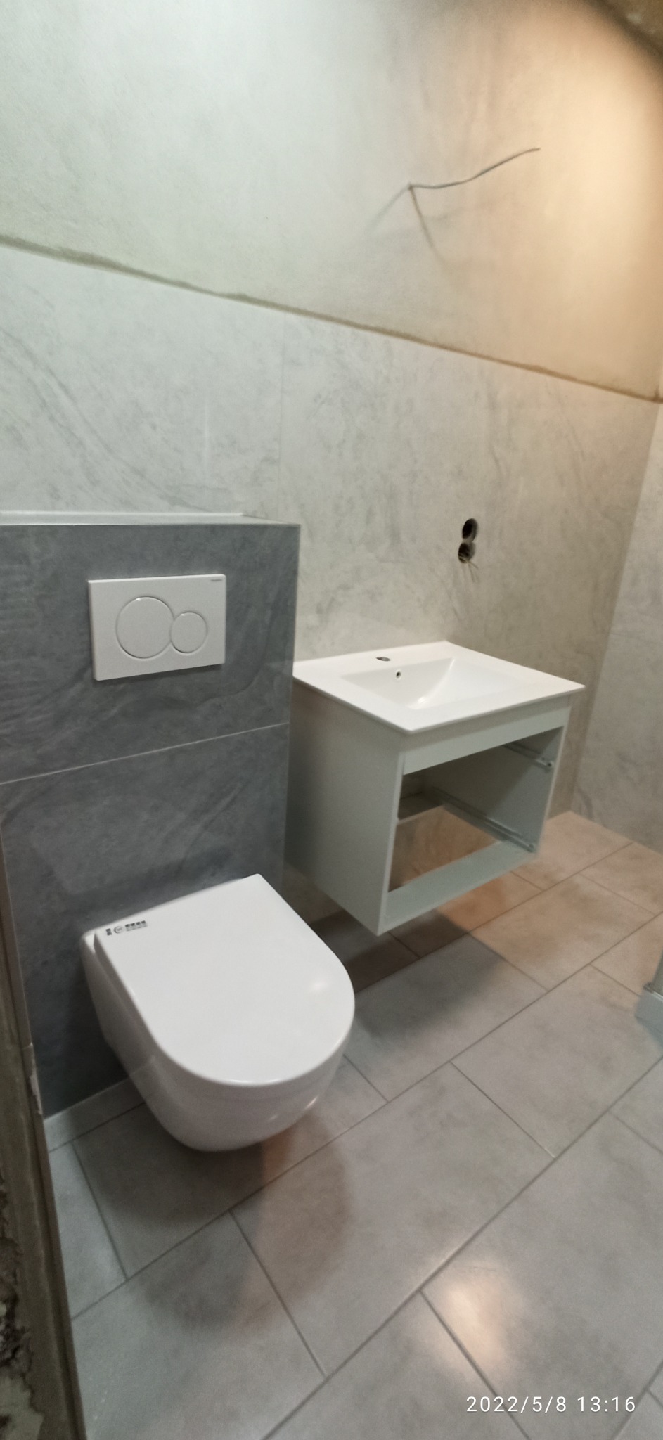 Wisząca toaleta i umywalka z szafką w surowym, wykafelkowanym wnętrzu łazienki z widocznymi kablami elektrycznymi w ścianie.