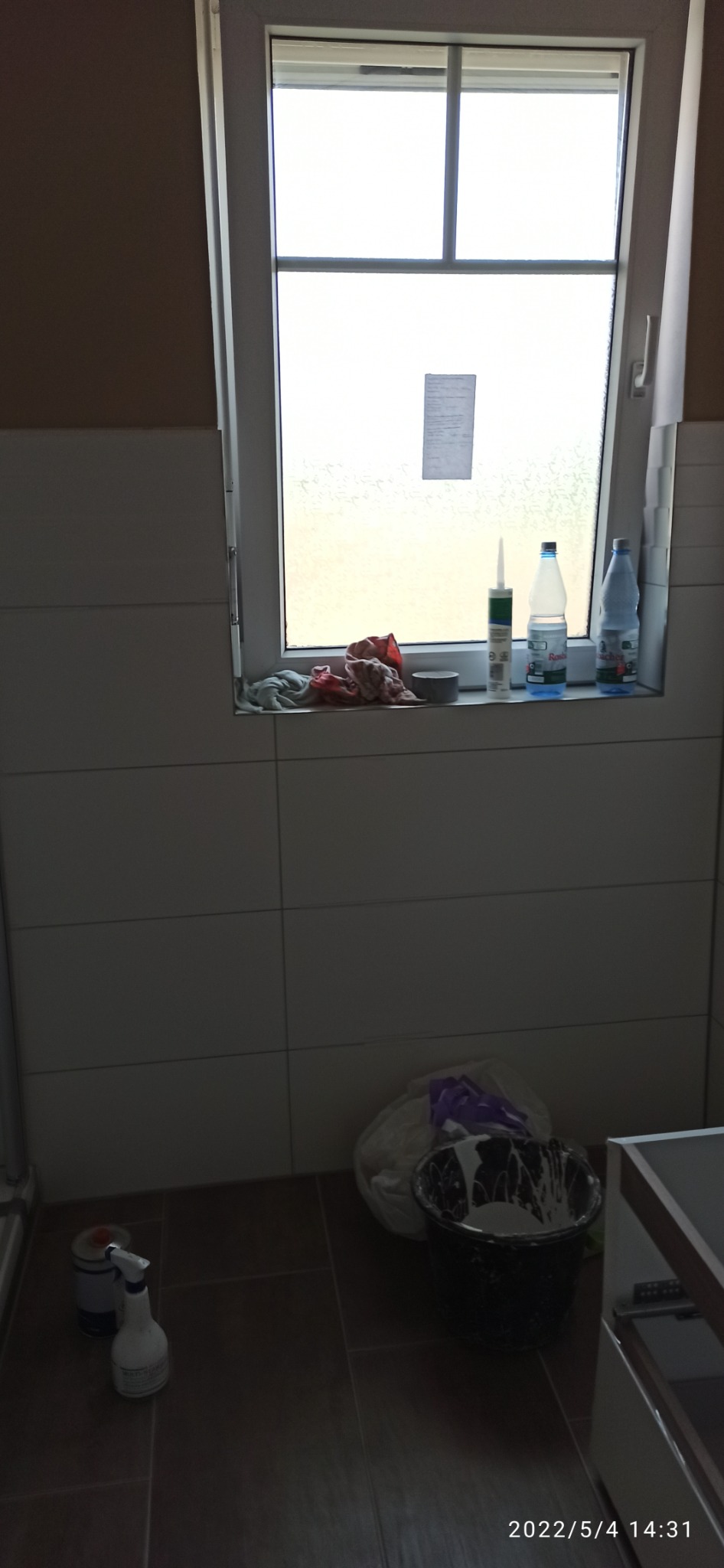 Remont łazienki: widok na okno z silikonem, butelkami i szmatkami na parapecie, poniżej wiadro z resztkami farby i środki czyszczące na podłodze z płytek.