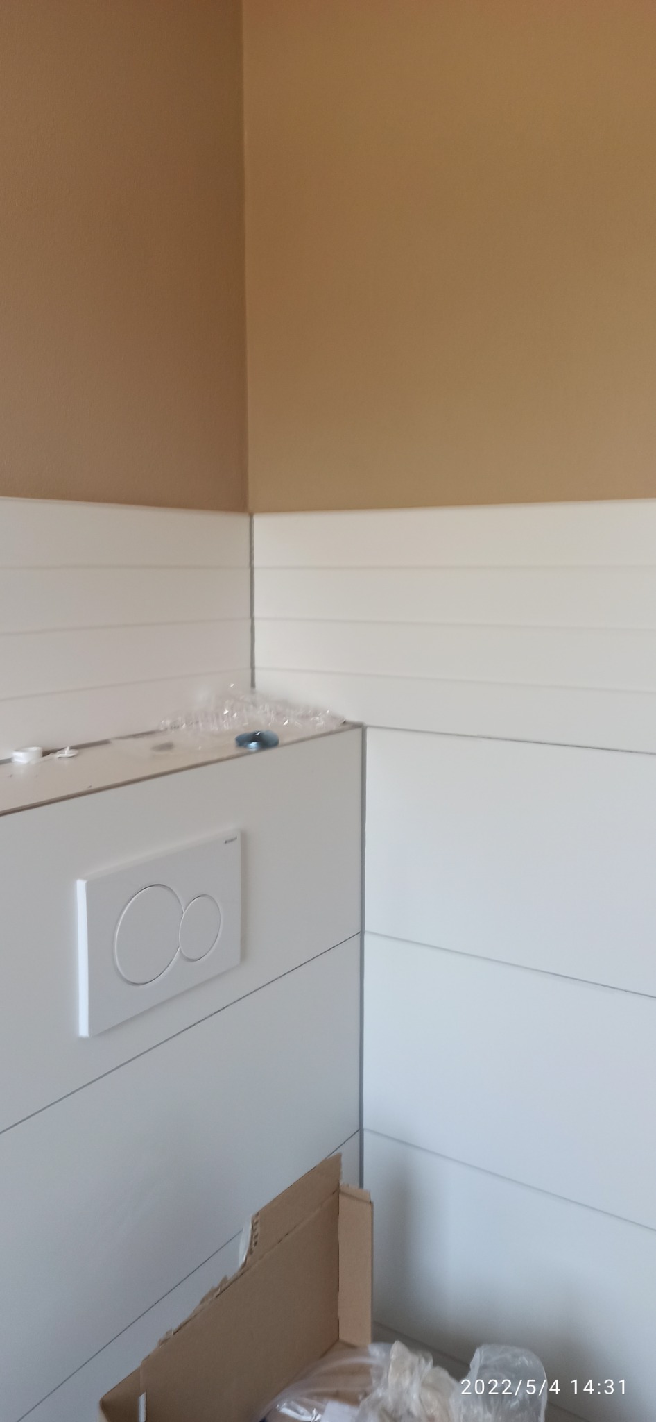 Świeżo zamontowany, podtynkowy stelaż WC z białym przyciskiem spłukującym na tle białej boazerii i beżowej ściany, widoczne ślady po montażu i karton po opakowaniu.