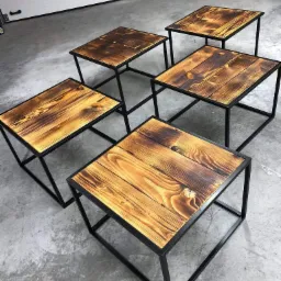 Zestaw pięciu identycznych, kwadratowych stolików kawowych z drewnianymi blatami o wyraźnym usłojeniu i metalowych, czarnych nogach, ustawionych na szarym, betonowym podłożu, z widoczną bramą...