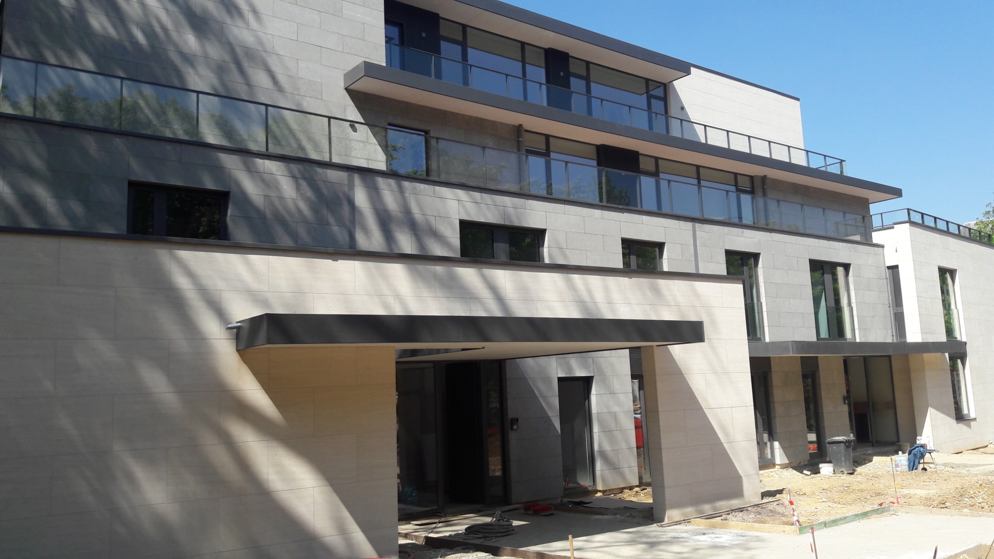 Nowoczesny budynek z jasnoszarą i ciemnoszarą elewacją z kamiennych płyt, widoczne okna z czarnymi ramami, balkony z szklanymi balustradami i zadaszone wejście, teren budowy przed budynkiem.