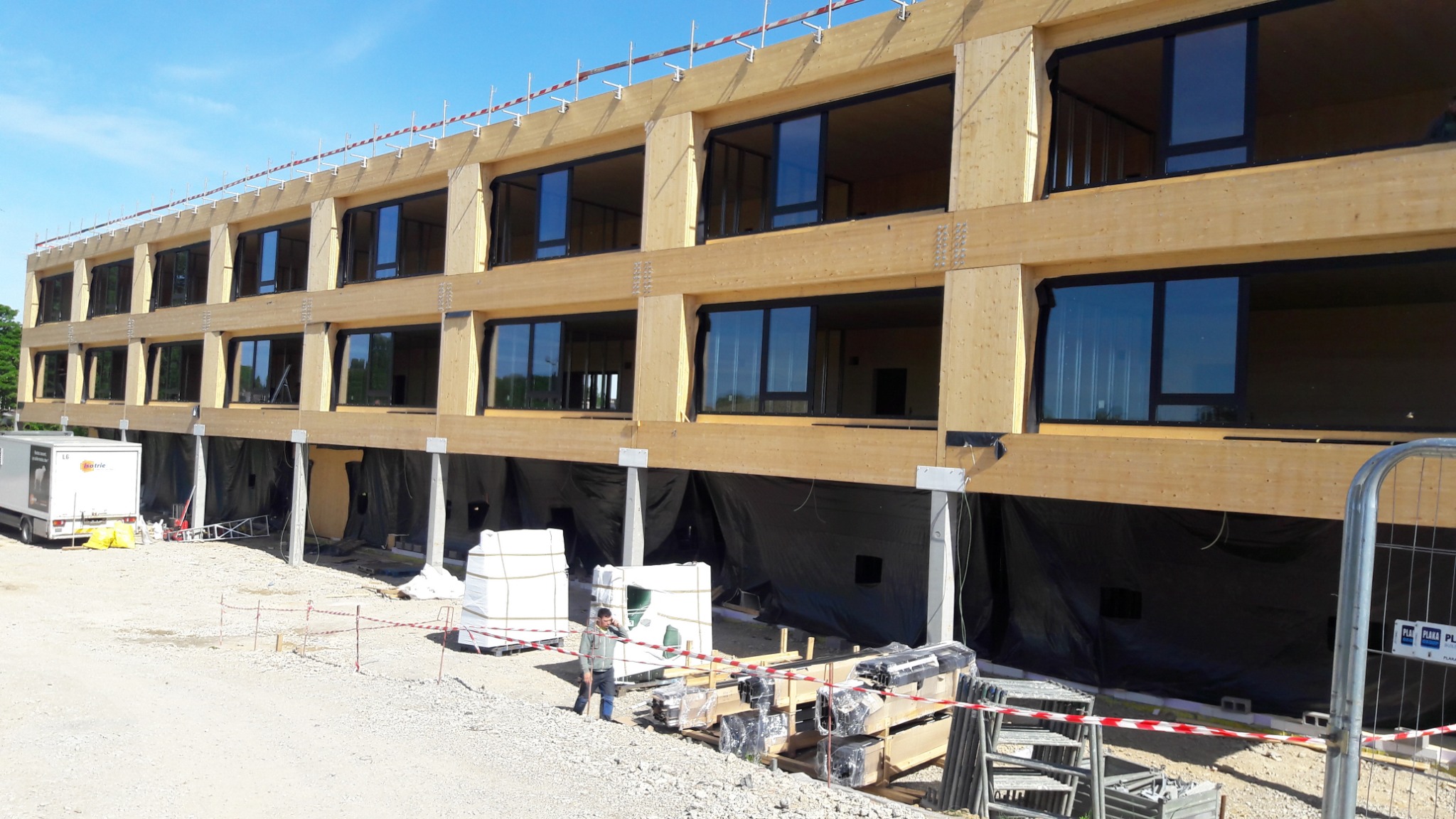 Wielopiętrowy budynek w trakcie budowy, z elementami drewnianymi i czarnymi ramami okiennymi, widoczne rusztowania, materiały budowlane i osoba w pobliżu, z lekkim pochyleniem kamery w lewo.