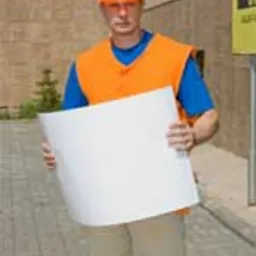 Mężczyzna w pomarańczowej kamizelce i kasku BHP trzymający białą kartkę papieru A3 na tle budynku z kostki brukowej i żółtego znaku informacyjnego.