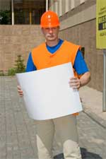 Mężczyzna w pomarańczowej kamizelce i kasku BHP trzymający białą kartkę papieru A3 na tle budynku z kostki brukowej i żółtego znaku informacyjnego.