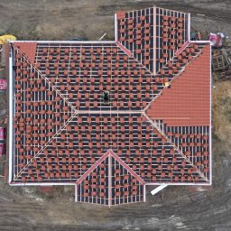 Nowakowski Twój Nowy Dach usługi dekarsko-budowlane Dariusz Nowakowski - Widok z góry na dach w trakcie krycia czerwoną dachówką. Część dachu już pokryta, reszta z widoczną konstrukcją i ułożonymi dachówkami gotowymi do montażu.