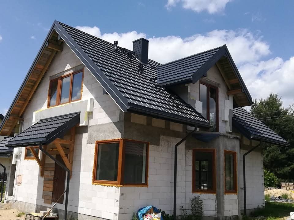 Dom w trakcie budowy z ciemną dachówką i drewnianą konstrukcją nad wejściem, widoczne okna z brązowymi ramami i fragment nieba z chmurami.