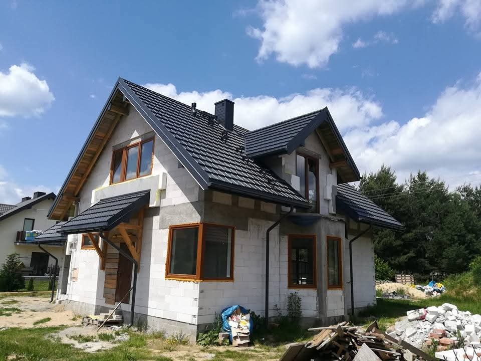 Dom jednorodzinny w trakcie budowy, z widocznym dachem pokrytym szarą dachówką, drewnianą konstrukcją nad wejściem i oknami w drewnianych ramach. W tle drzewa i błękitne niebo.