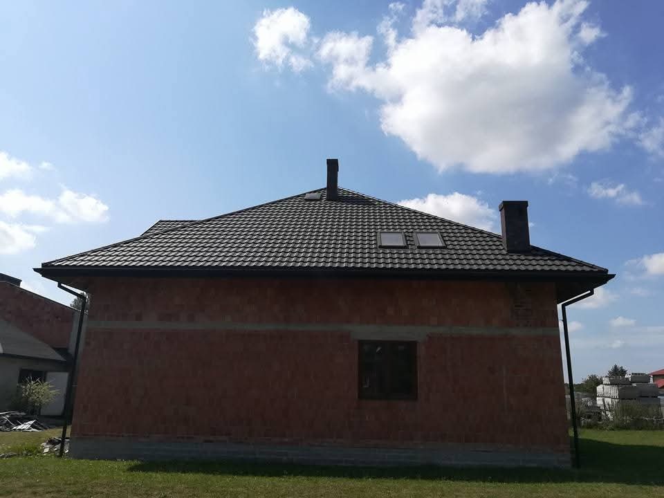 Dom w trakcie budowy z czarnym dachem z dachówki, widoczne okna dachowe i rynna. Ściany z czerwonej cegły, błękitne niebo z chmurami w tle.