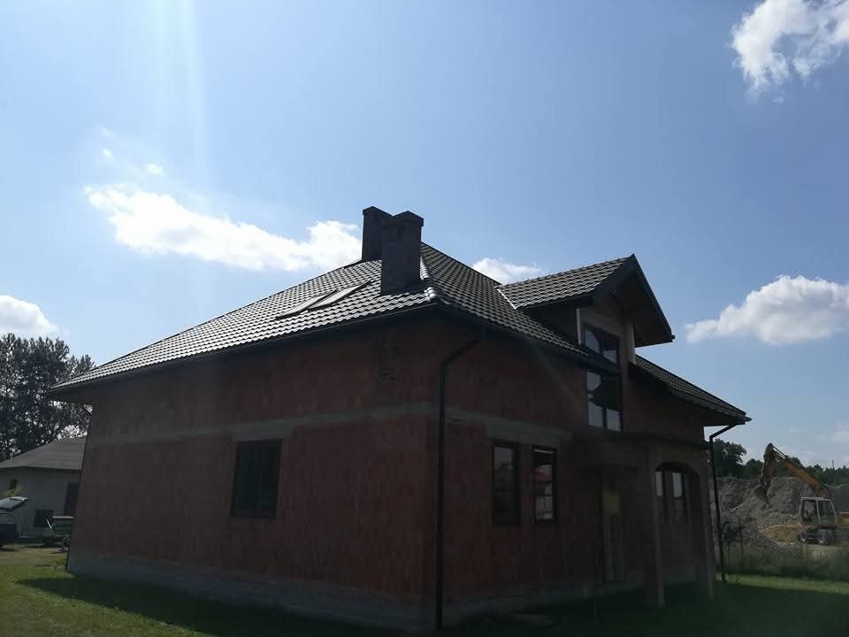 Dom w trakcie budowy z ciemnym dachem z dachówki, widoczne okno dachowe i rynny. W tle błękitne niebo z chmurami i koparka.