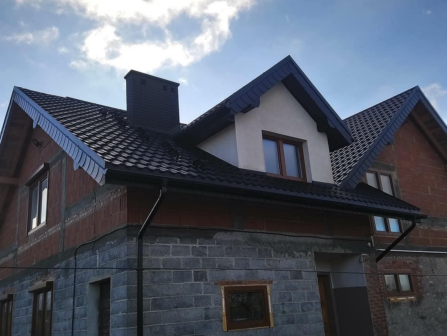 Dom w trakcie budowy z czarnym dachem z blachy, widoczne rynny i komin. Część budynku w stanie surowym, okna zamontowane. Pochmurne niebo w tle.