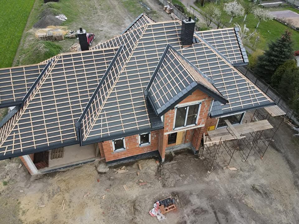 Dom w trakcie krycia dachu z czarną dachówką, widoczne łaty, komin i rusztowania. Ujęcie z drona ukazuje postęp prac na tle zieleni.