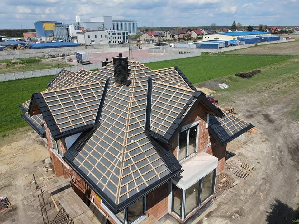 Dom w trakcie krycia dachu z czarną dachówką, widoczna konstrukcja drewniana, komin z cegły klinkierowej, w tle zabudowa wiejska i pole uprawne.