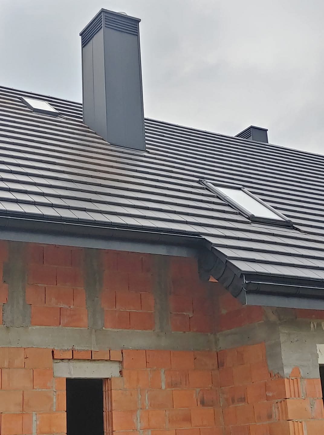 Nowy dach z ciemnej dachówki ceramicznej, z oknami dachowymi i kominem wentylacyjnym. Widoczny fragment muru z czerwonej cegły i orynnowanie.