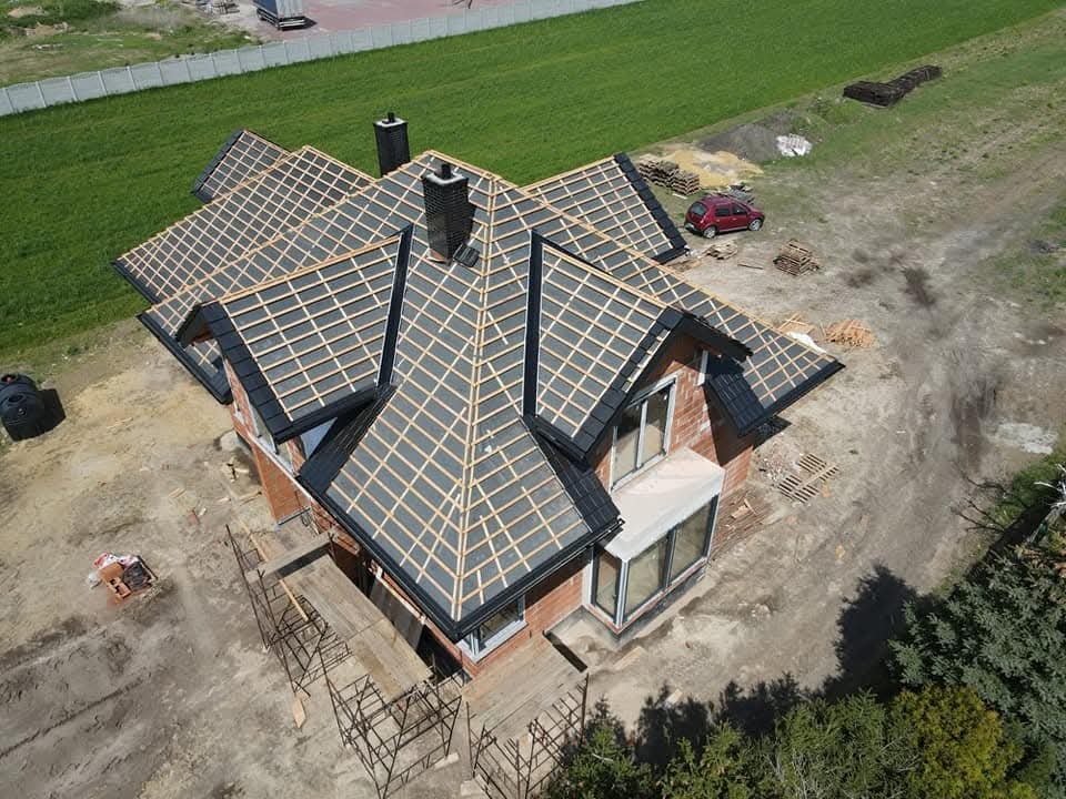 Dom w trakcie krycia dachu z czarną dachówką, widok z góry ukazuje konstrukcję więźby i częściowo ułożone pokrycie. Na działce widoczne materiały budowlane.
