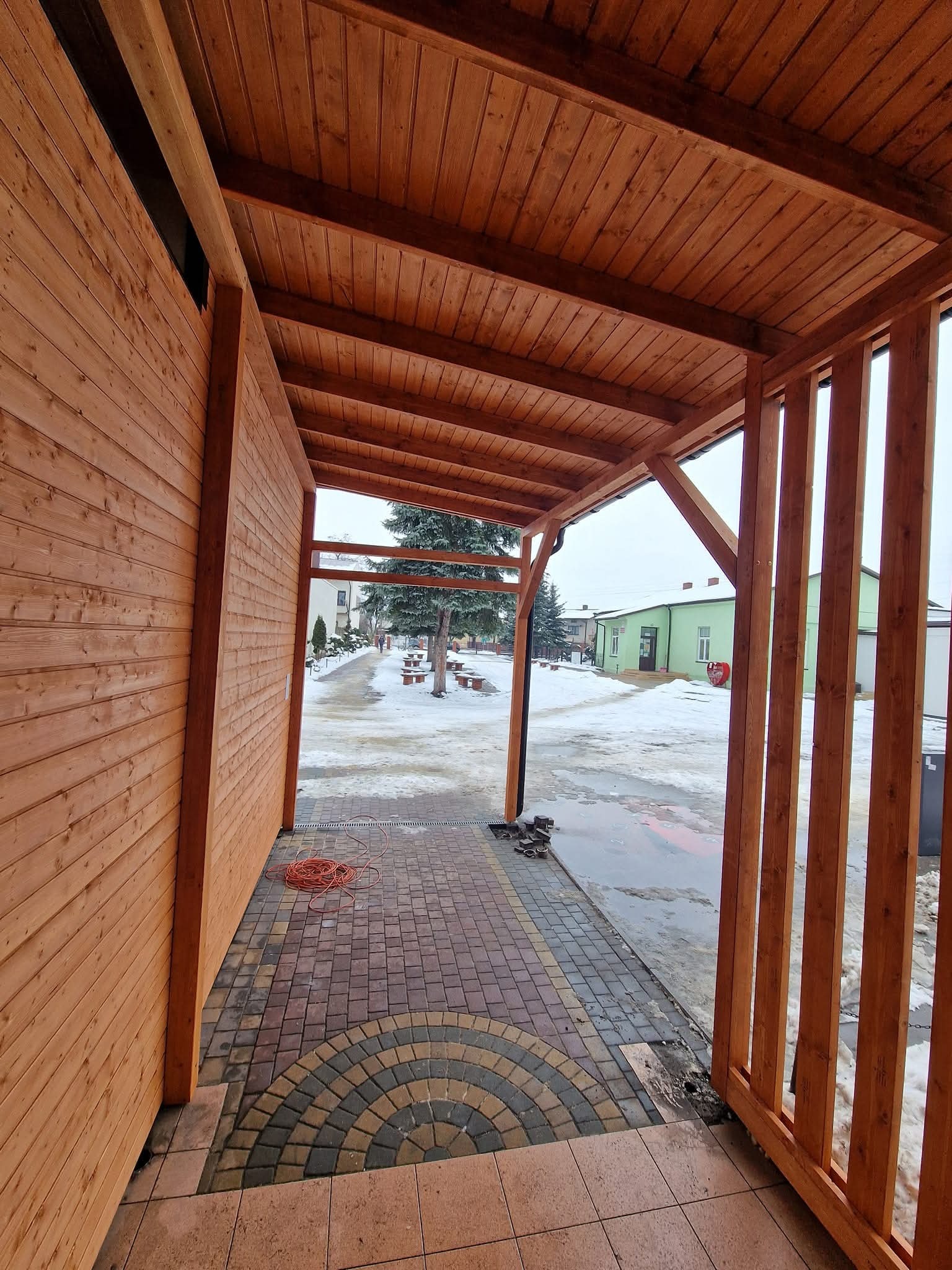 Drewniana wiata z widokiem na zimowy krajobraz. Konstrukcja z belek, zadaszenie z desek, dekoracyjna kostka brukowa. Widoczny kabel elektryczny.