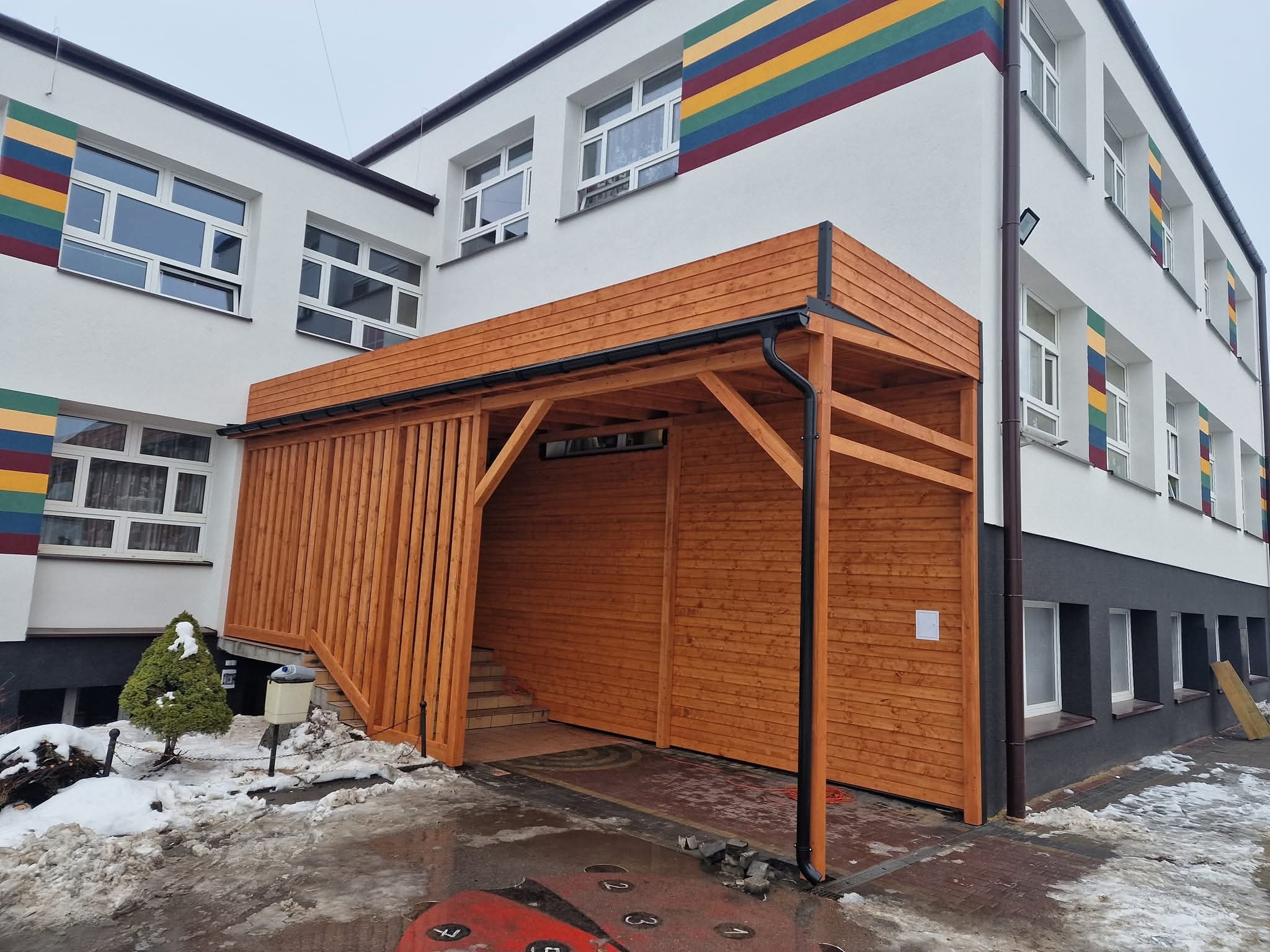 Drewniana wiata z rynnami przy budynku szkolnym z kolorowymi pasami. Widoczne schody wejściowe i częściowo ośnieżony teren.