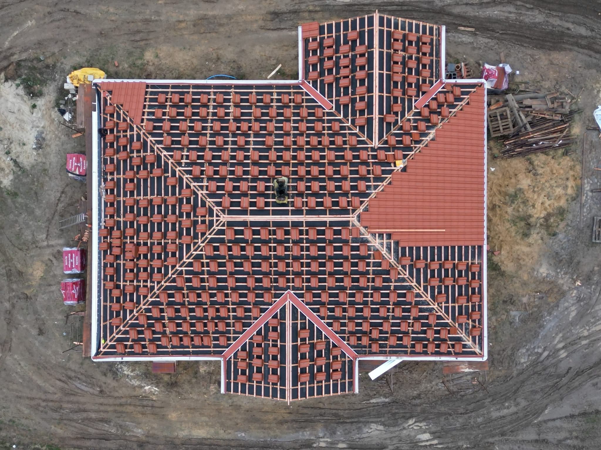 Widok z góry na dach w trakcie krycia czerwoną dachówką. Część dachu już pokryta, reszta z widoczną konstrukcją i ułożonymi dachówkami gotowymi do montażu.