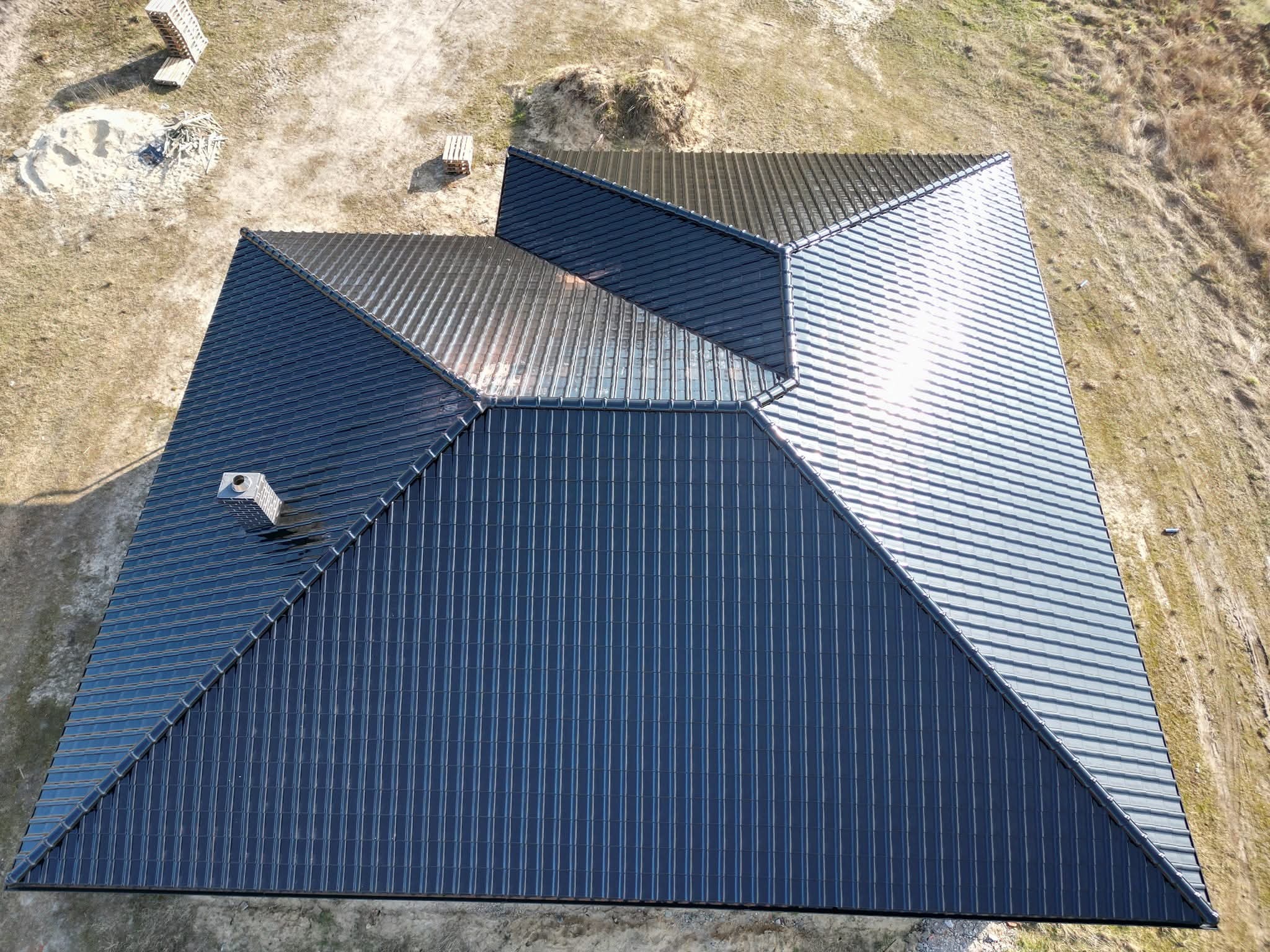 Nowoczesny, ciemny dach z blachy na domu jednorodzinnym, widok z góry. Charakterystyczny komin i symetryczny układ połaci. W tle teren budowy z paletami.