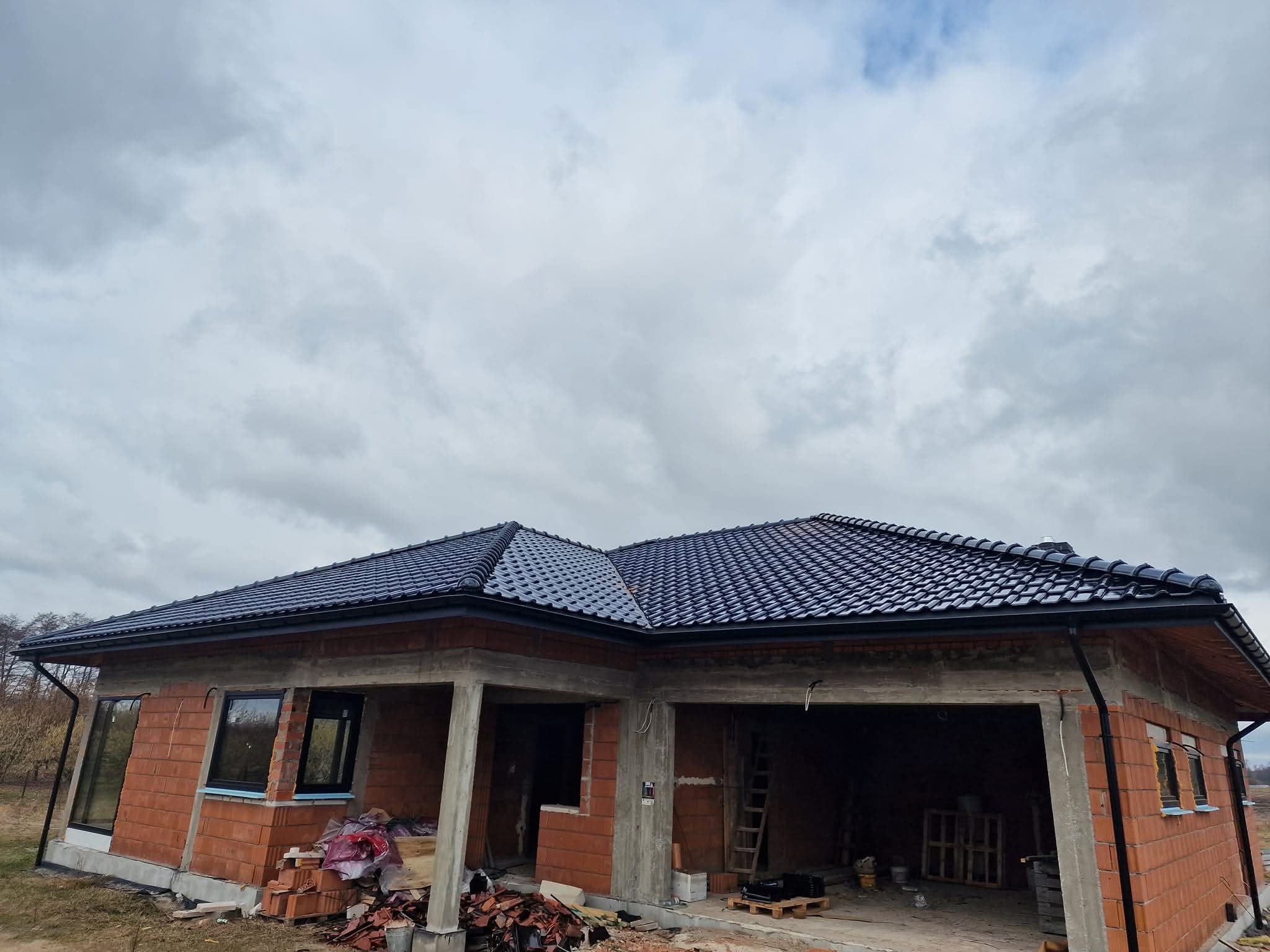 Dom w trakcie budowy z ciemną dachówką i zamontowanymi rynnami, widoczne cegły i betonowe elementy konstrukcyjne, pochmurne niebo w tle.