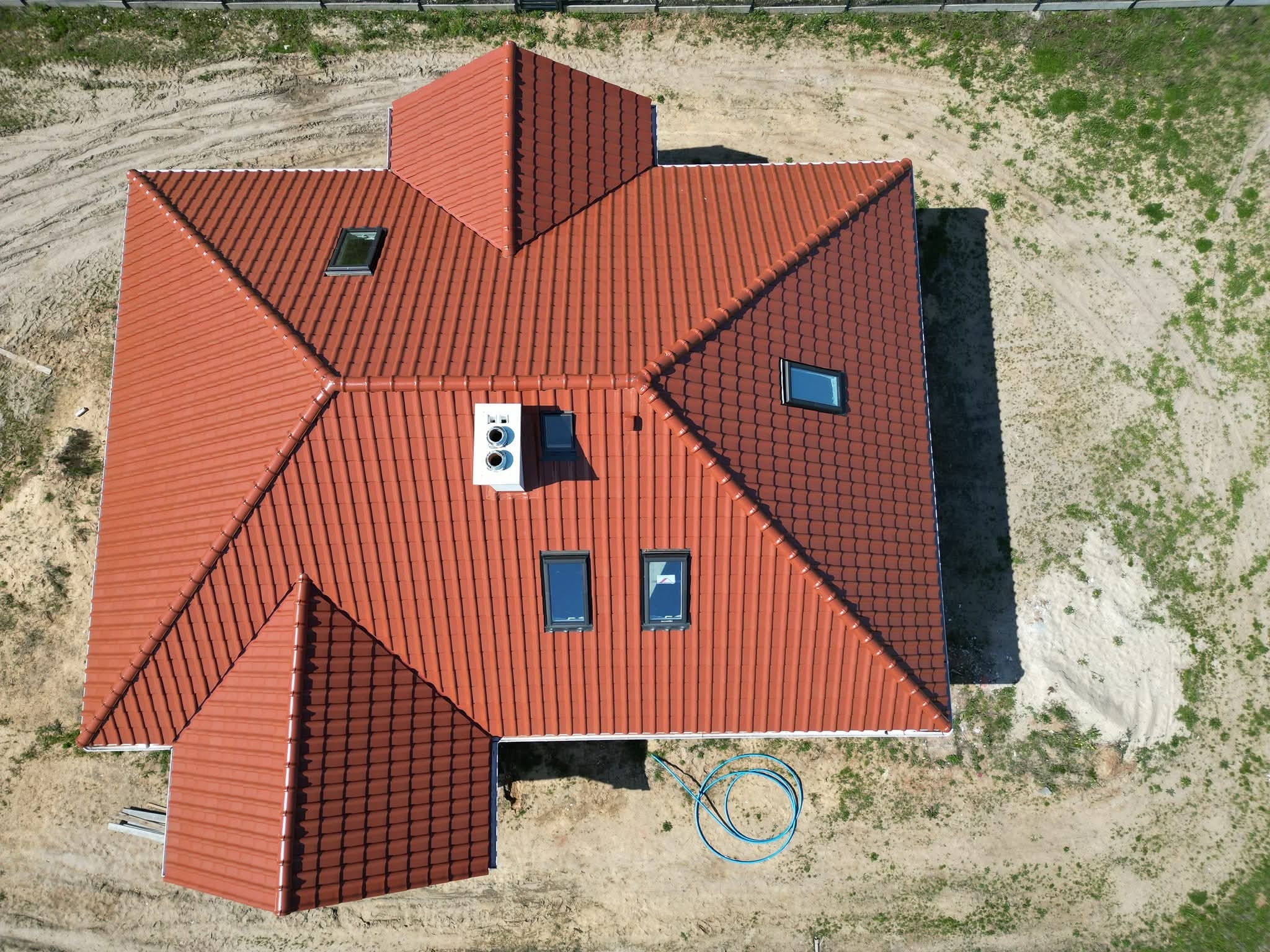 Widok z góry na dom z czerwoną dachówką, oknami dachowymi i kominem. Na ziemi leży zwinięty niebieski wąż. Ujęcie z drona.