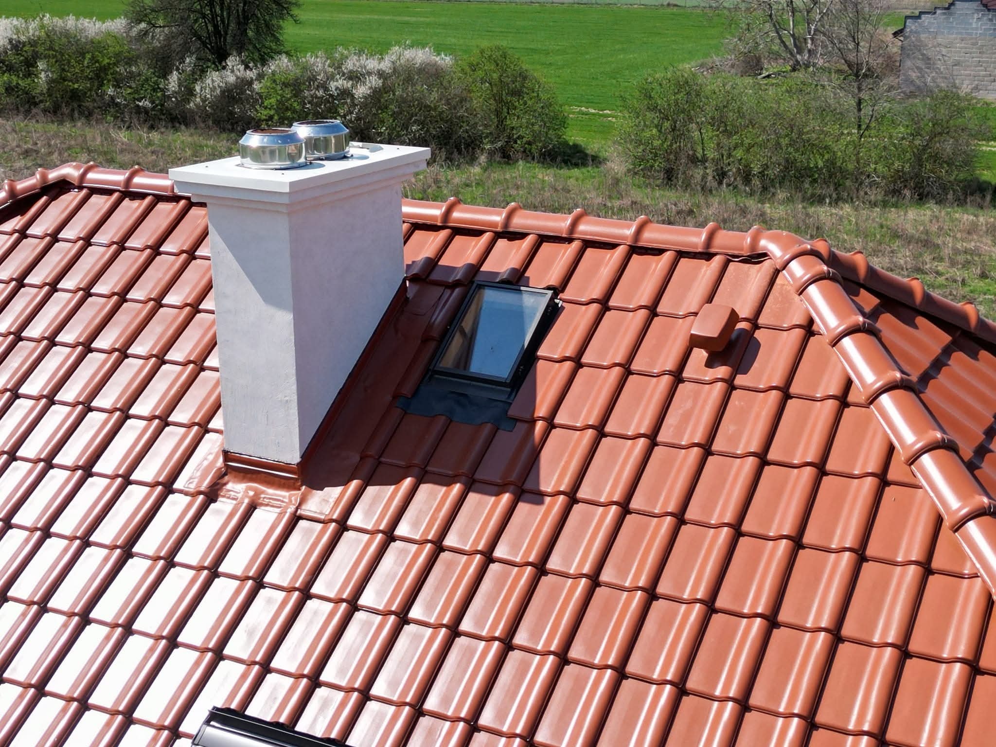 Czerwona dachówka ceramiczna z kominem i oknem dachowym, widok z góry na tle zieleni. Nowoczesne pokrycie dachowe z detalami.