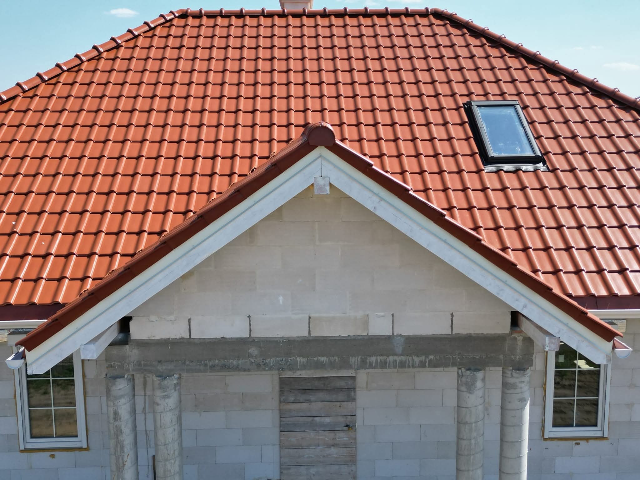 Czerwony dach z dachówką ceramiczną i oknem dachowym na budynku w trakcie budowy, widoczne białe ściany z betonu komórkowego i fragment okna.