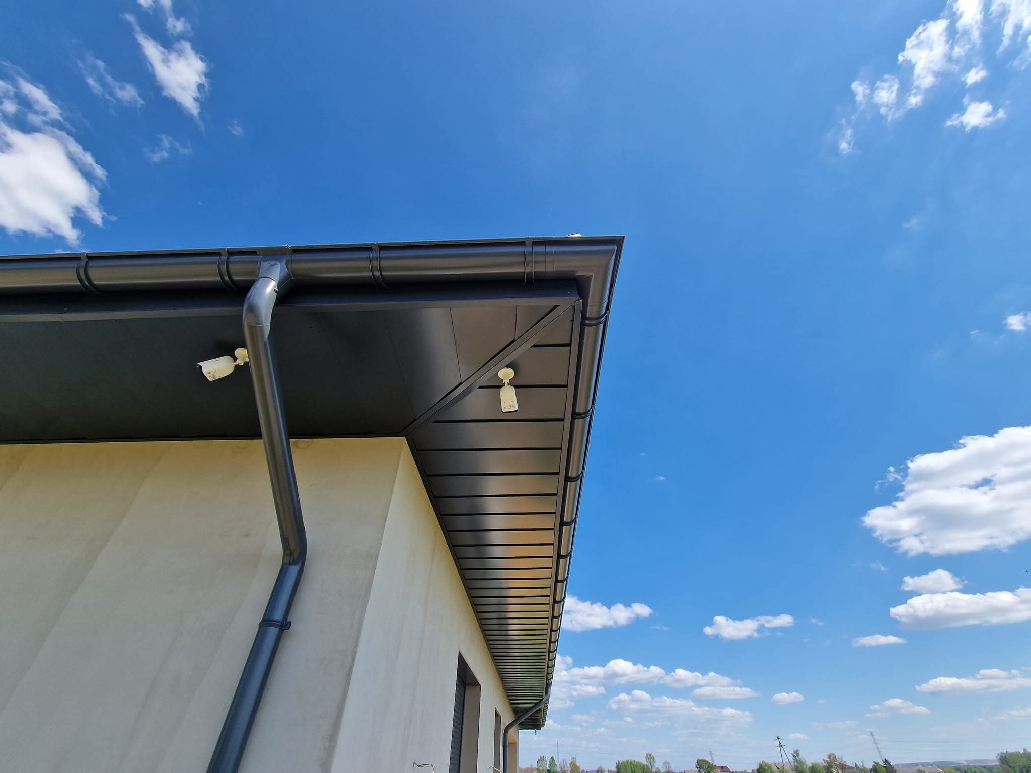 Nowoczesny, grafitowy dach z blachy na jasnym budynku, widok z dołu na tle błękitnego nieba z chmurami. Widoczne rynny i kamery.