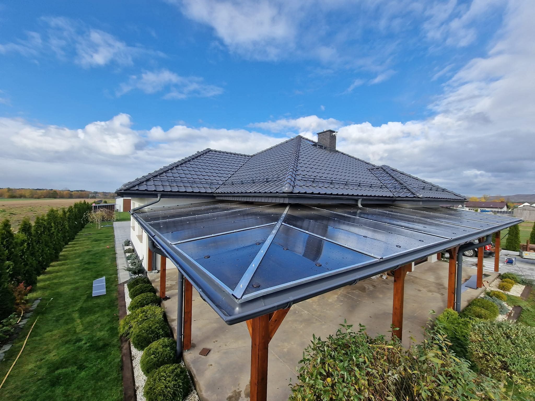 Zadaszenie tarasu z poliwęglanu trapezowego, wsparte na drewnianych słupach, z widocznym dachem domu krytym dachówką ceramiczną w tle, w słoneczny dzień.