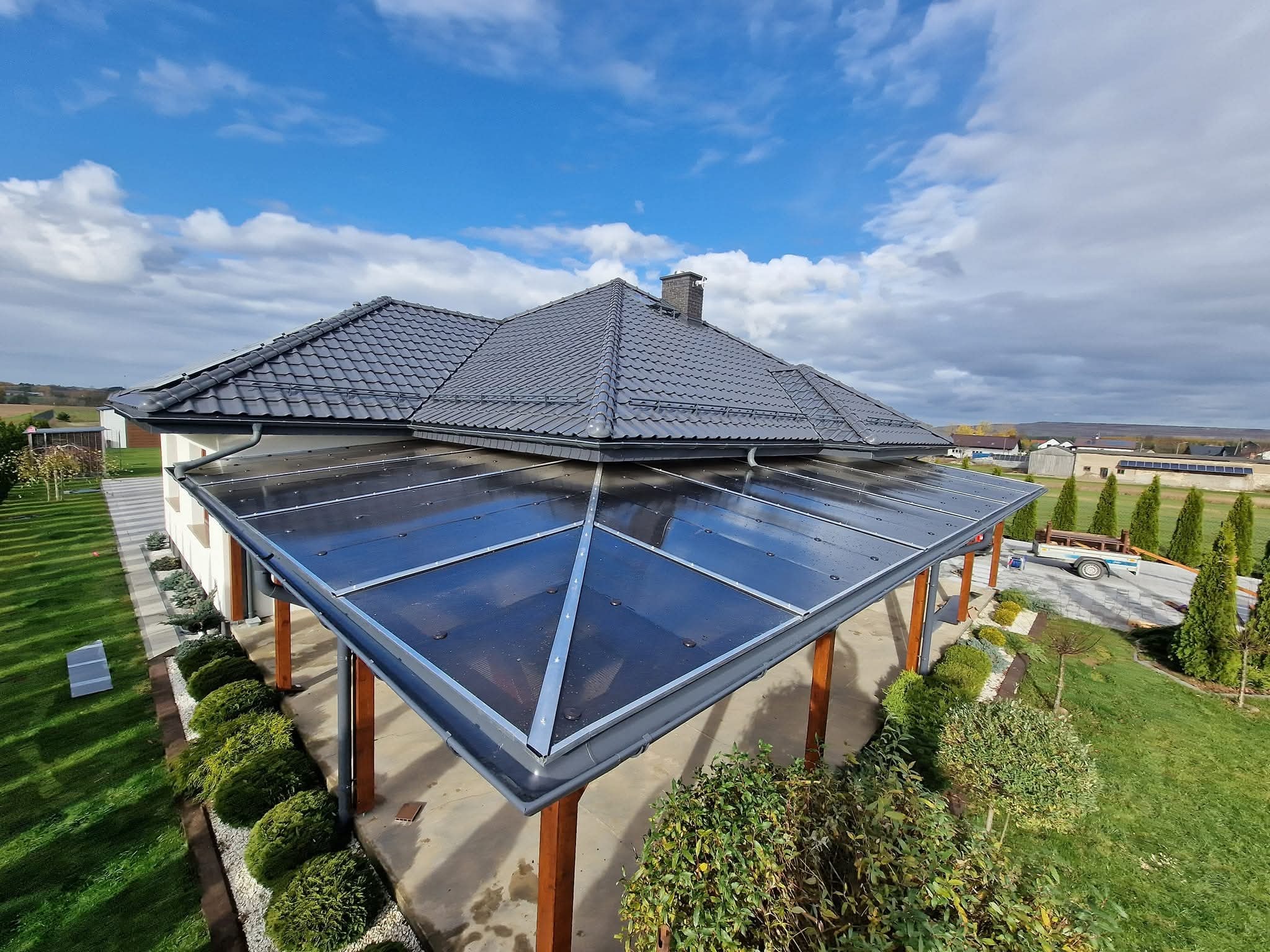 Dom z ciemną dachówką i zadaszeniem tarasu z przezroczystych paneli, widok z góry na tle błękitnego nieba z chmurami. Widoczne rynny i elementy konstrukcyjne.