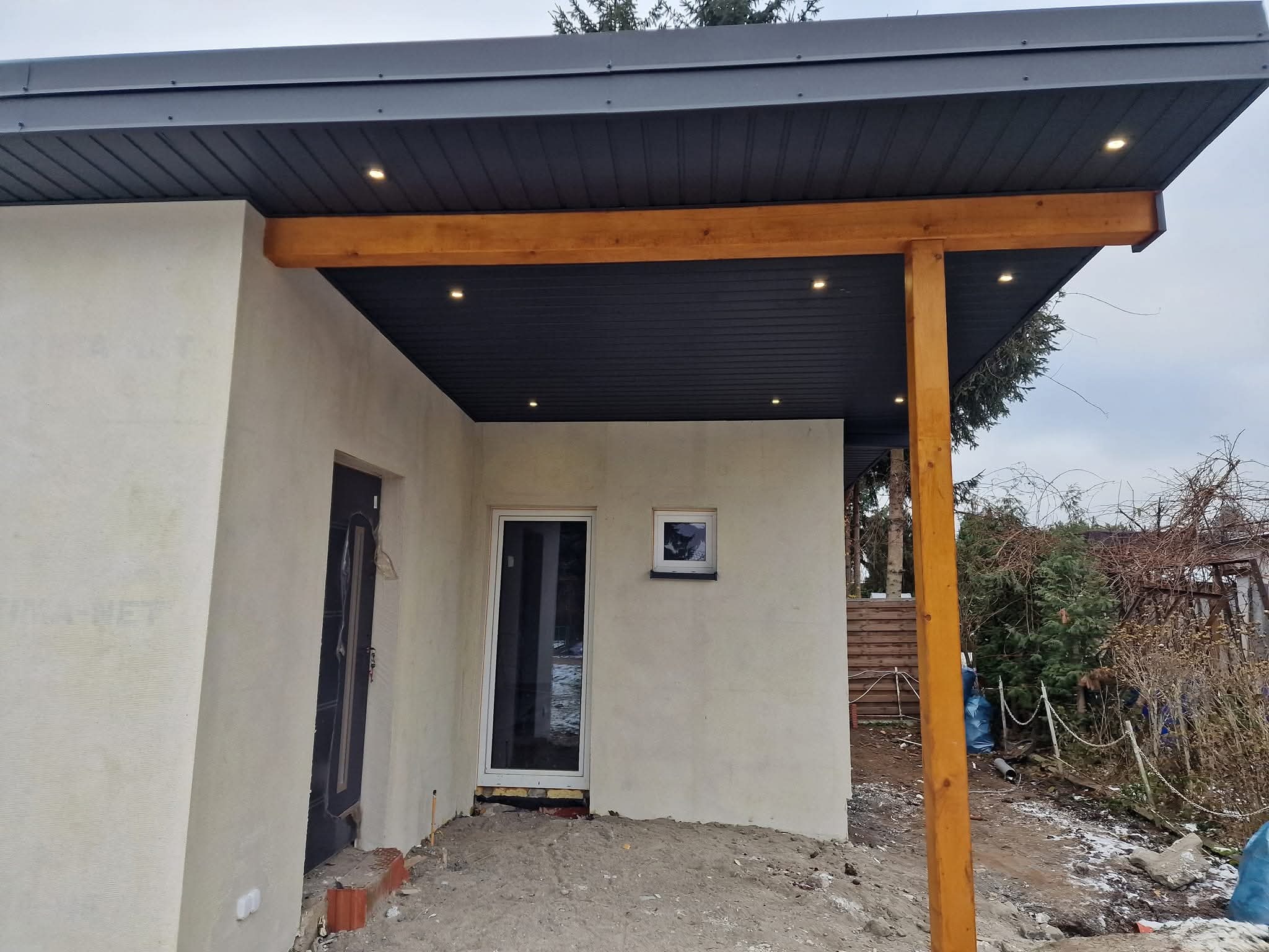 Zadaszenie tarasu z drewnianą konstrukcją i oświetleniem punktowym w Rusiec. Nowoczesne wykończenie elewacji budynku z drzwiami i oknami.