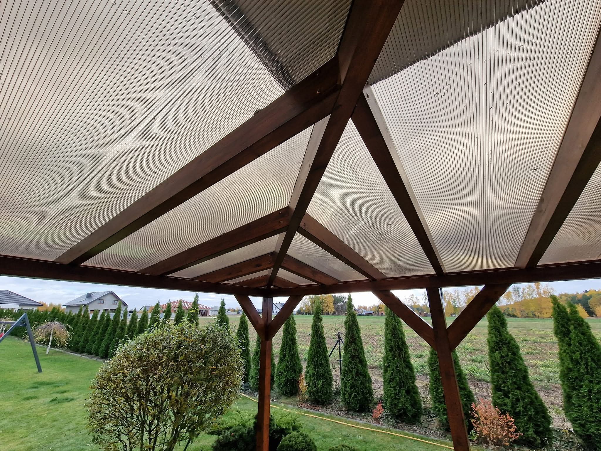 Zadaszenie tarasu z brązową drewnianą konstrukcją i przezroczystym, żebrowanym pokryciem, widok z dołu na tle zieleni ogrodu i zabudowań.