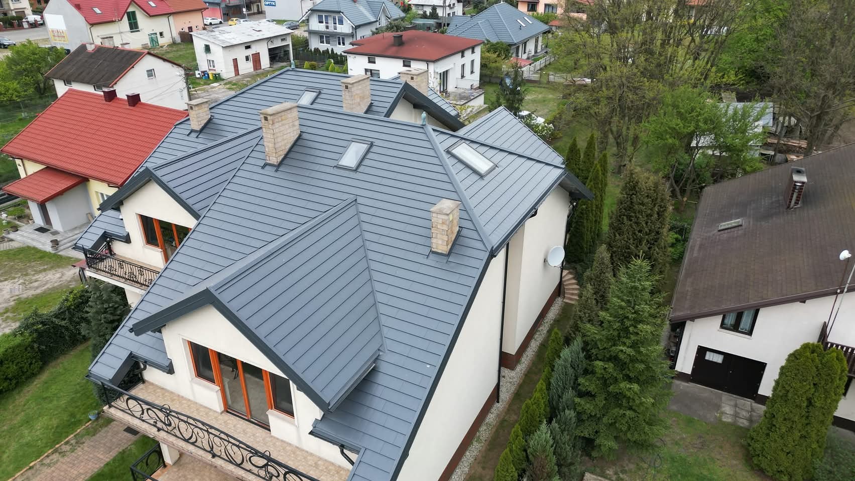 Dom z szarym dachem z blachy na rąbek stojący, widok z góry. Na dachu okna połaciowe i kominy z cegły. W tle inne domy i zieleń.