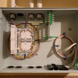 Otwarta skrzynka rozdzielcza z kablami światłowodowymi ułożonymi wewnątrz, widoczne złącza, adaptery i zapas kabla. Przewody uporządkowane za pomocą niebieskich uchwytów. Na dnie skrzynki cztery...