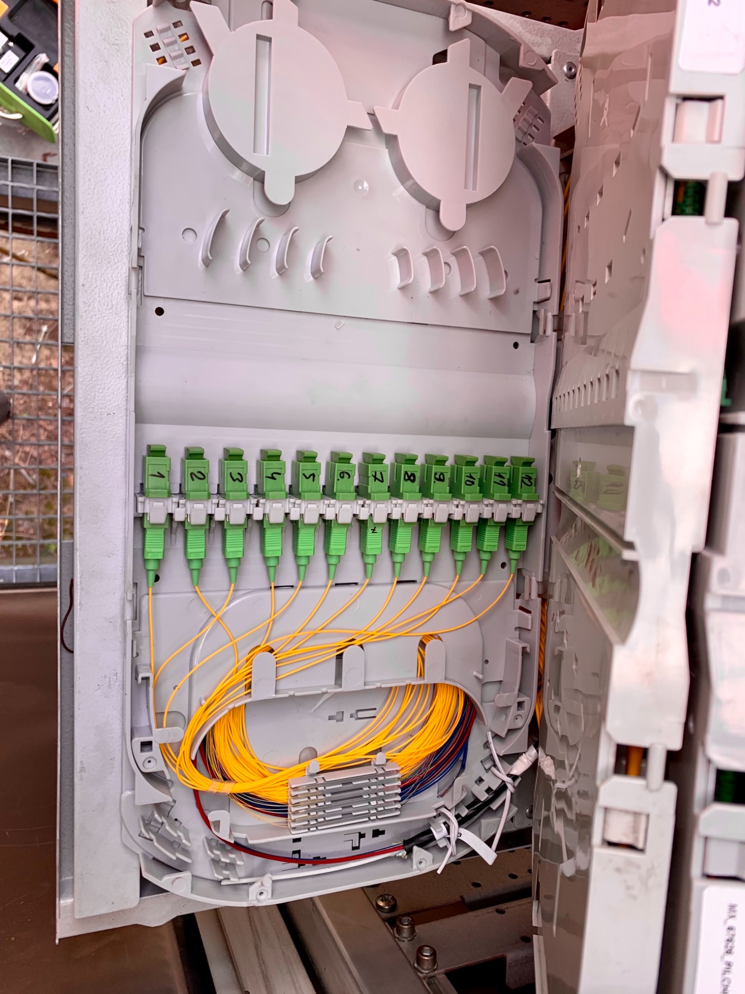 Otwarta skrzynka telekomunikacyjna z podłączonymi światłowodami oznaczonymi numerami od 1 do 12, widoczne żółte kable światłowodowe i złącza w kolorze zielonym.